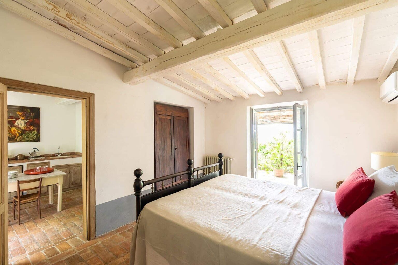 Cozy bedroom with a double bed, featuring red accent pillows and a wooden ceiling. The room includes an open window with natural light and a view of greenery, along with a doorway leading to a kitchen area. Ideal for a relaxing stay. | Only on SopranoVillas