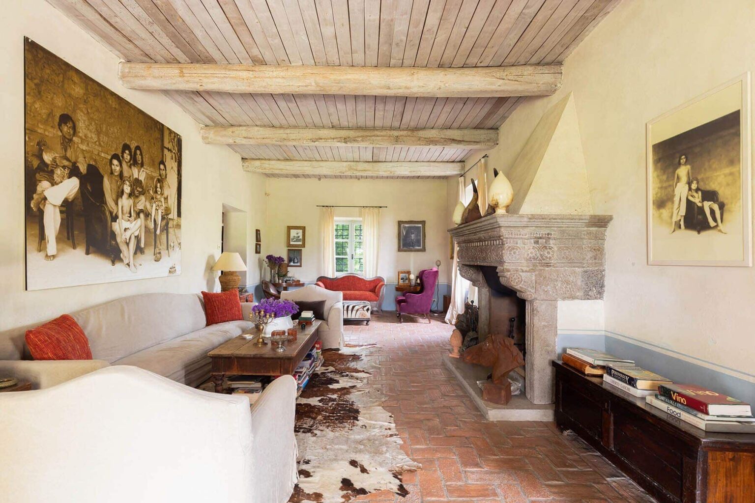 Spacious and inviting living room with rustic wooden beams, featuring a cozy fireplace and eclectic artwork on the walls. The room is adorned with comfortable seating, including a large sofa, vibrant cushions, and stylish decor elements, creating a warm and welcoming atmosphere. | Only on SopranoVillas