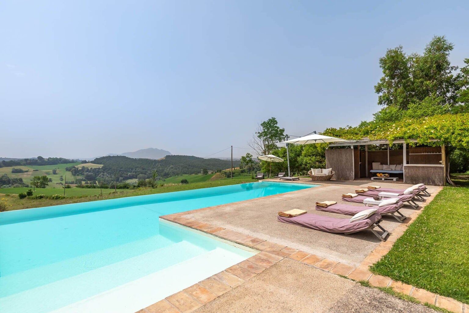 Luxury villa with a stunning infinity pool overlooking lush green hills and a serene landscape, featuring sun loungers and a shaded seating area. Ideal for a relaxing getaway. | Only on SopranoVillas
