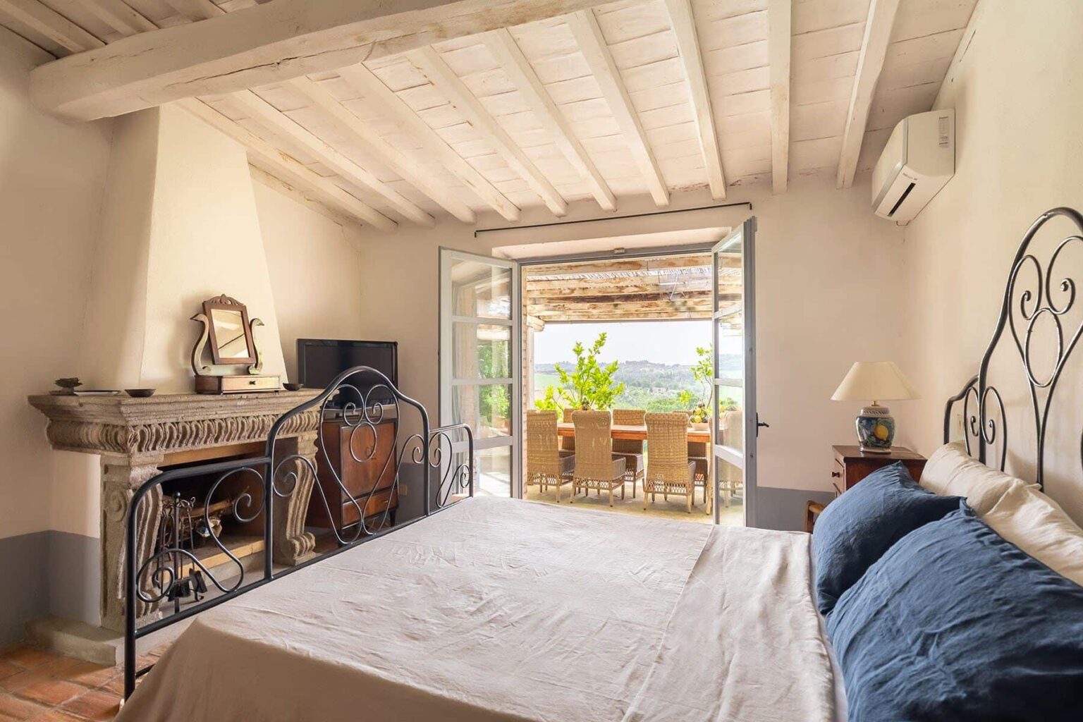 Cozy bedroom with a wrought iron bed, featuring a fireplace and a view of an outdoor dining area through large sliding glass doors, enhancing the bright and airy atmosphere. | Only on SopranoVillas