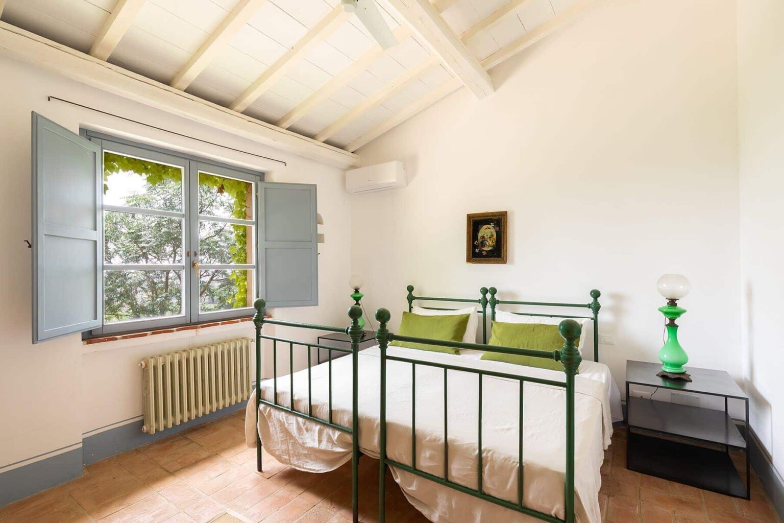 Cozy bedroom featuring a wrought iron bed with green accents, large windows with wooden shutters, and a vintage bedside lamp. The light-colored walls and wooden ceiling create a warm, inviting atmosphere, ideal for relaxation. | Only on SopranoVillas