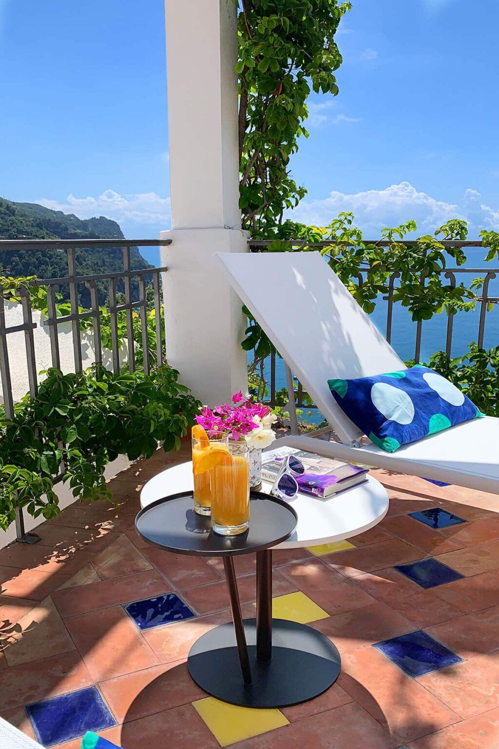 Lounge chair on a balcony overlooking a scenic view, with a table featuring colorful cocktails, fresh flowers, and a stack of magazines. Ideal for relaxation and enjoying outdoor living. | Only on SopranoVillas