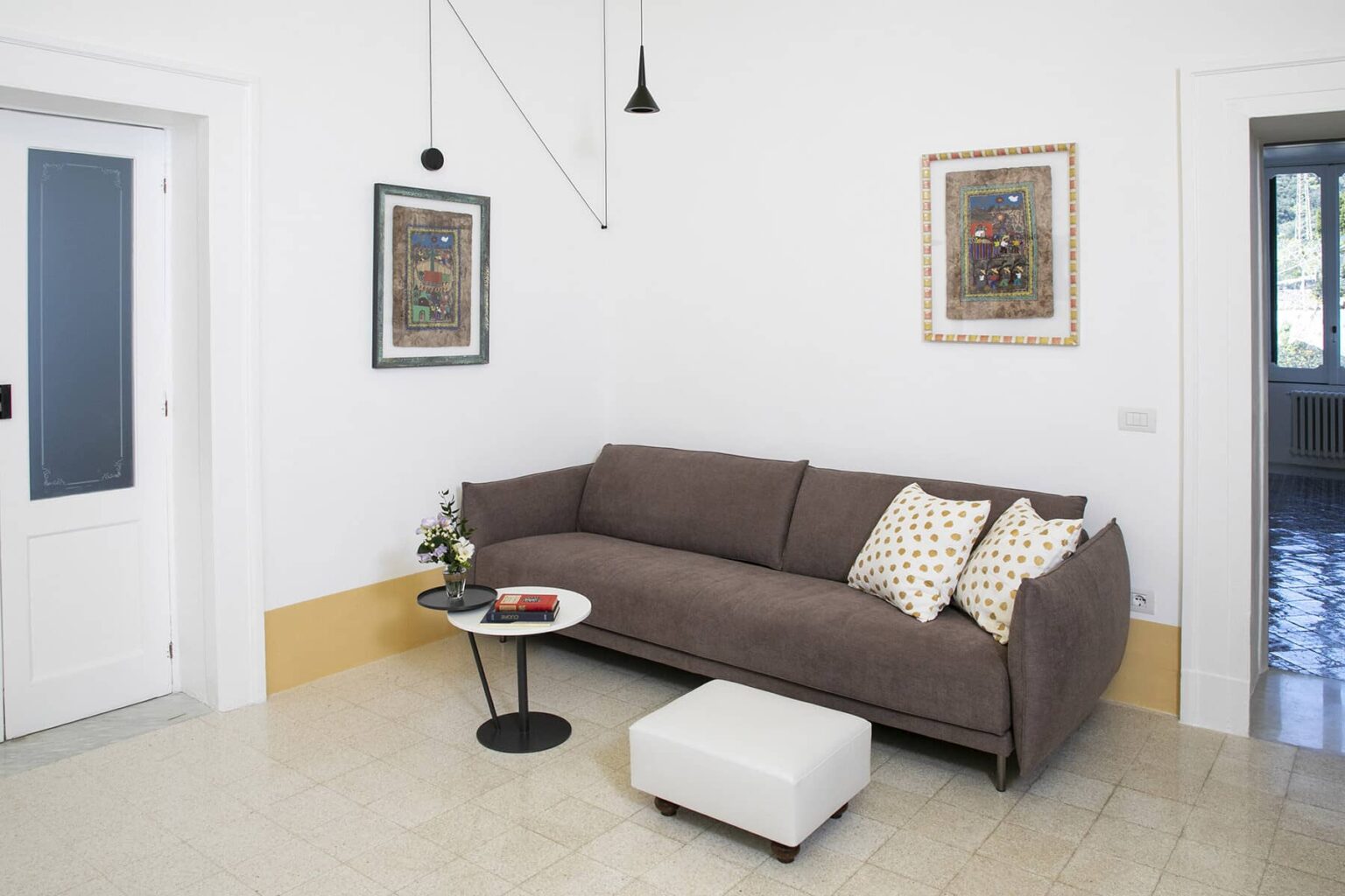 Modern living room featuring a cozy brown sofa with decorative pillows, a minimalist black side table with a book, and a white ottoman. The room has white walls, framed artwork, and natural light coming through a nearby door. | Only on SopranoVillas