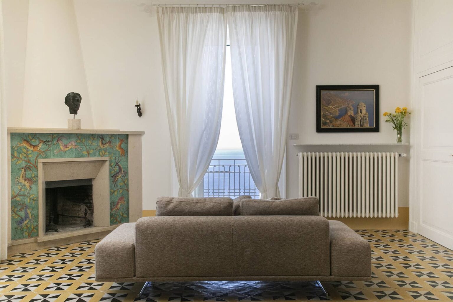 Modern living room featuring a beige sofa, decorative fireplace with colorful tiles, large windows with sheer curtains, and a painting on the wall, creating a bright and inviting atmosphere. | Only on SopranoVillas
