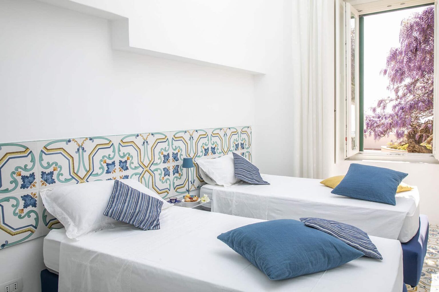 Bright and airy bedroom featuring two twin beds with blue decorative pillows, a colorful tiled headboard, and a window showcasing a view of blooming purple flowers outside. | Only on SopranoVillas