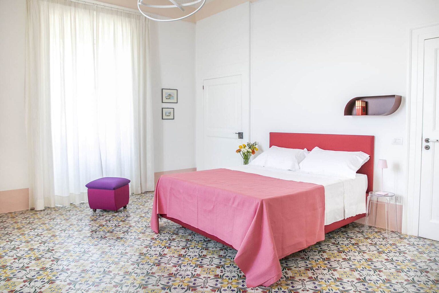 Bright and airy bedroom featuring a red bed with white bedding, a pink throw blanket, and a small purple ottoman. The room has large windows with sheer curtains, allowing natural light to illuminate the colorful patterned floor tiles. A minimalist shelf displays decorative items, enhancing the modern aesthetic. | Only on SopranoVillas