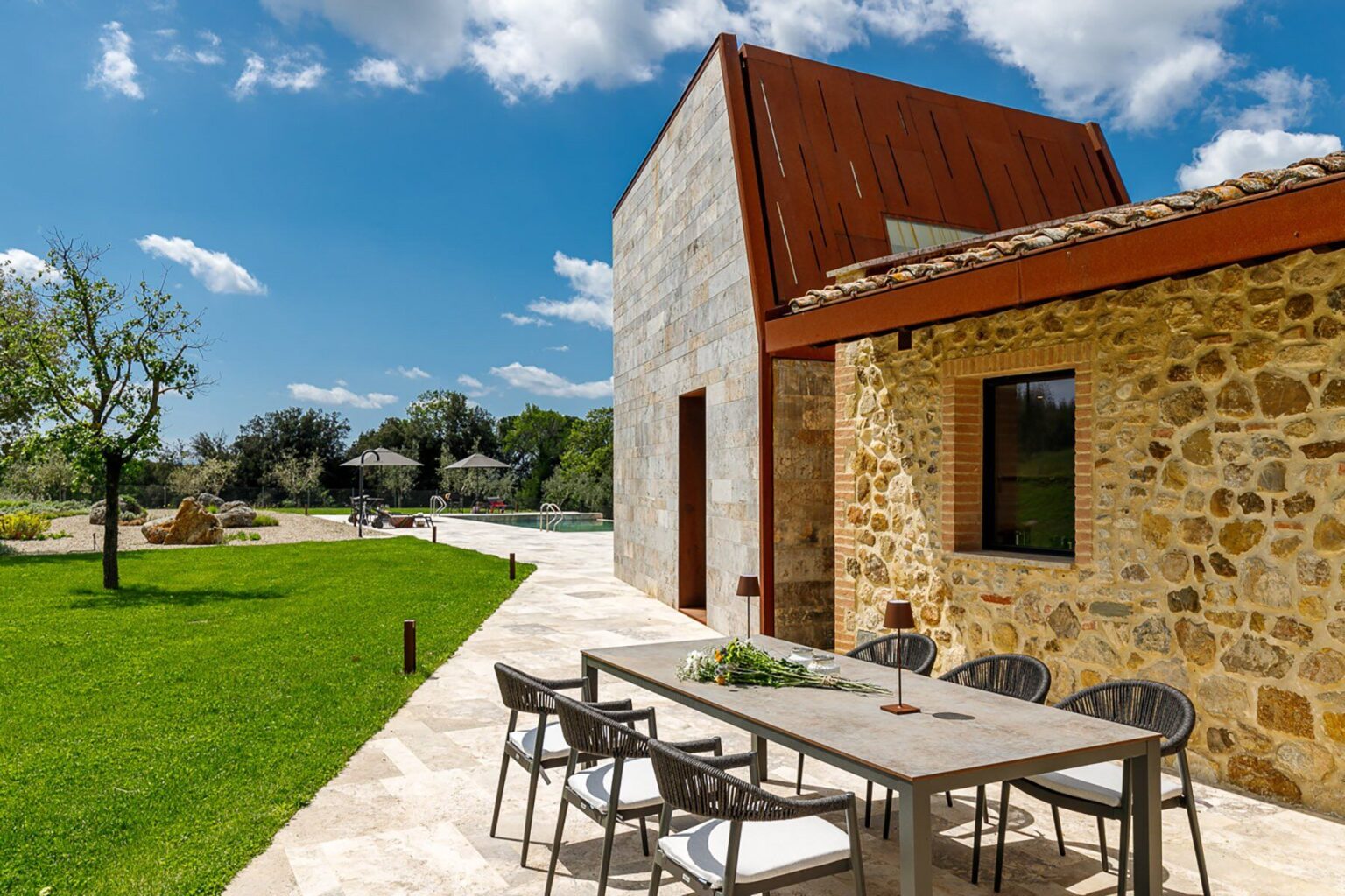 Modern stone house with a rustic design, featuring a spacious patio with outdoor seating, surrounded by lush green grass and trees, under a bright blue sky. | Only on SopranoVillas