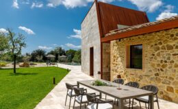 Modern stone house with a rustic design, featuring a spacious patio with outdoor seating, surrounded by lush green grass and trees, under a bright blue sky.