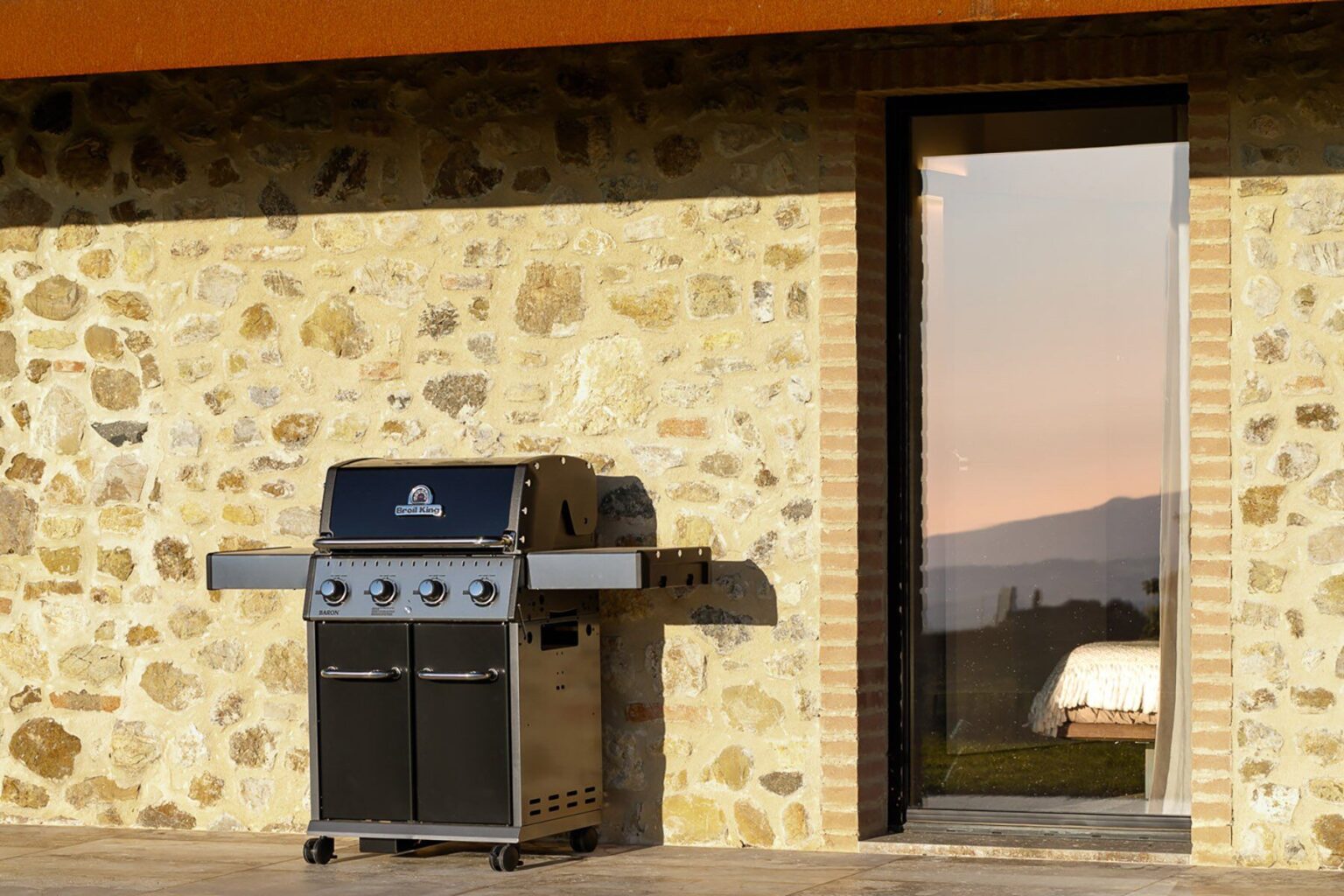 Modern gas grill on a stone patio with a large glass door, overlooking a scenic outdoor view at sunset. Perfect for outdoor cooking and entertaining. | Only on SopranoVillas
