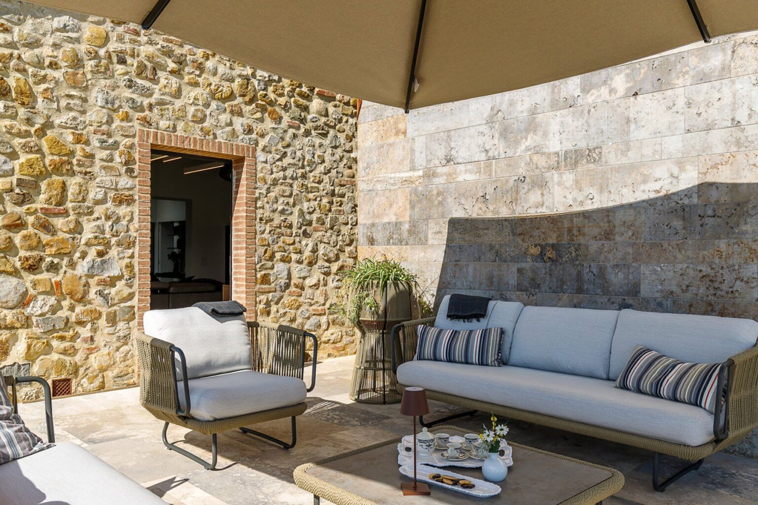 Cozy outdoor seating area featuring a stylish sofa with cushions and a coffee table, set against a rustic stone wall and shaded by a large canopy. Perfect for relaxation and entertaining in a serene environment. | Only on SopranoVillas