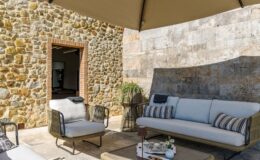 Cozy outdoor seating area featuring a stylish sofa with cushions and a coffee table, set against a rustic stone wall and shaded by a large canopy. Perfect for relaxation and entertaining in a serene environment.