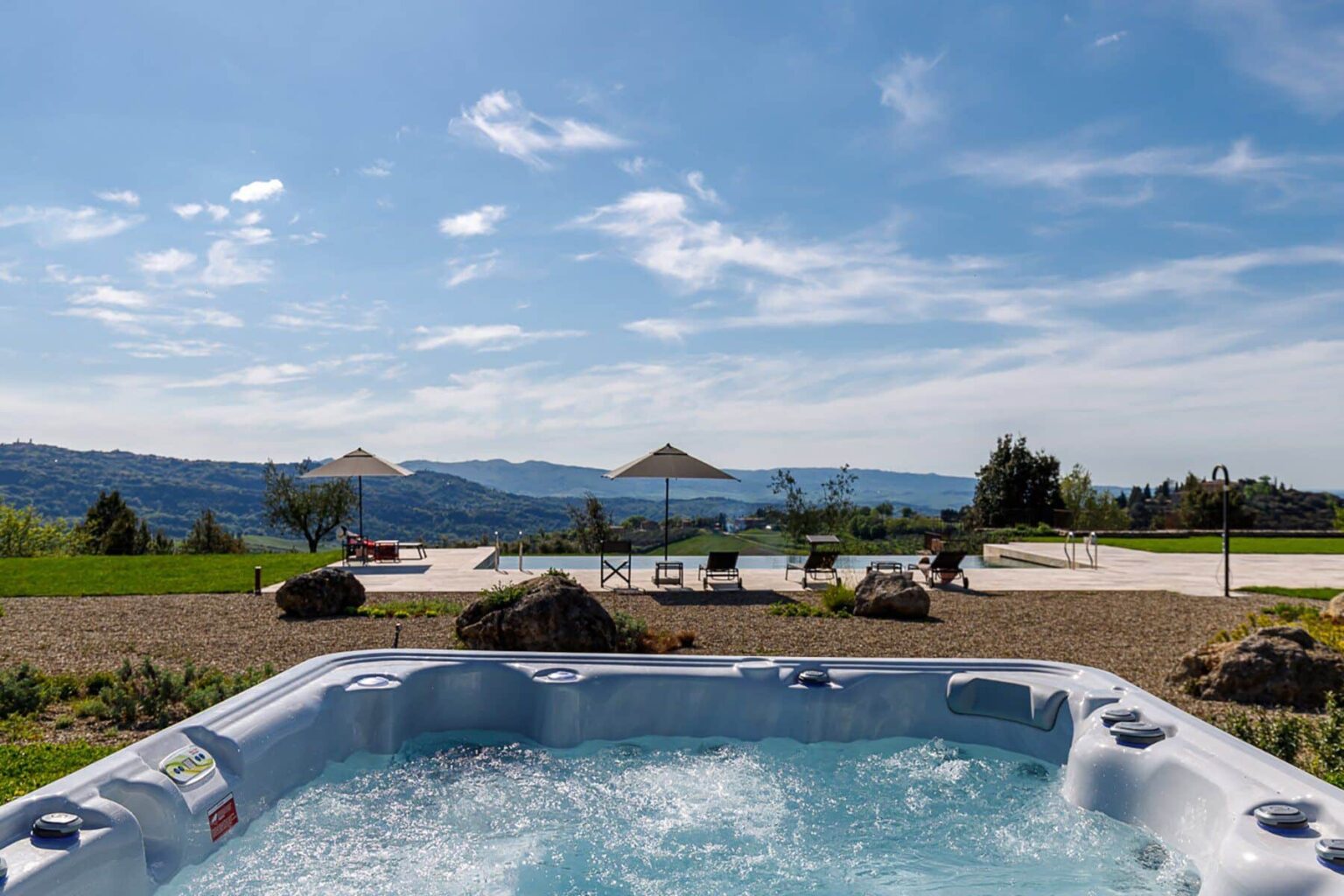 Hot tub overlooking scenic landscape with mountains and blue sky, featuring lounge chairs and umbrellas in a serene outdoor setting. | Only on SopranoVillas