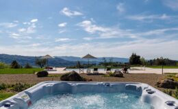 Hot tub overlooking scenic landscape with mountains and blue sky, featuring lounge chairs and umbrellas in a serene outdoor setting.