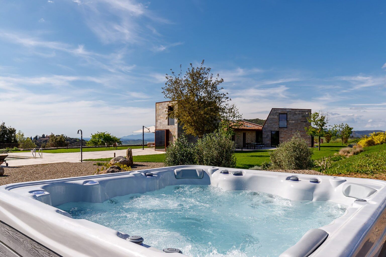 Relaxing outdoor hot tub with bubbling water, surrounded by lush greenery and a scenic view of a modern house and landscape under a clear blue sky. Perfect for leisure and wellness. | Only on SopranoVillas