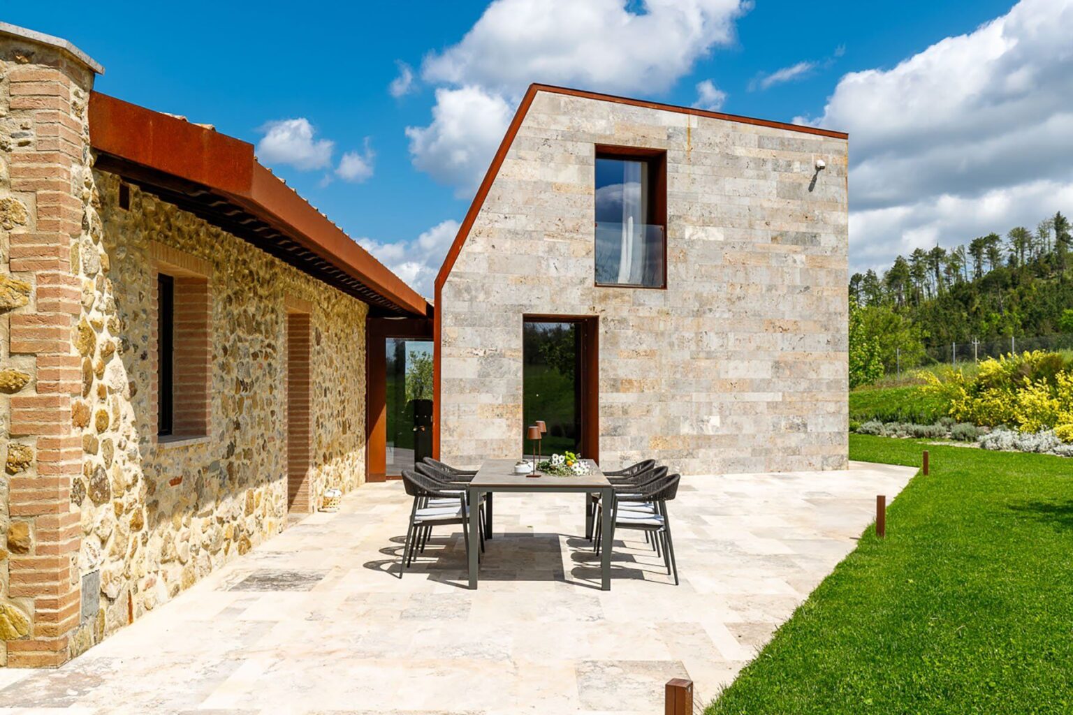 Modern stone house with a unique angular design, featuring a patio area with outdoor dining furniture, surrounded by lush greenery and blue skies. Ideal for showcasing contemporary architecture and outdoor living spaces. | Only on SopranoVillas