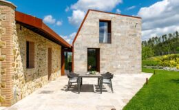Modern stone house with a unique angular design, featuring a patio area with outdoor dining furniture, surrounded by lush greenery and blue skies. Ideal for showcasing contemporary architecture and outdoor living spaces.