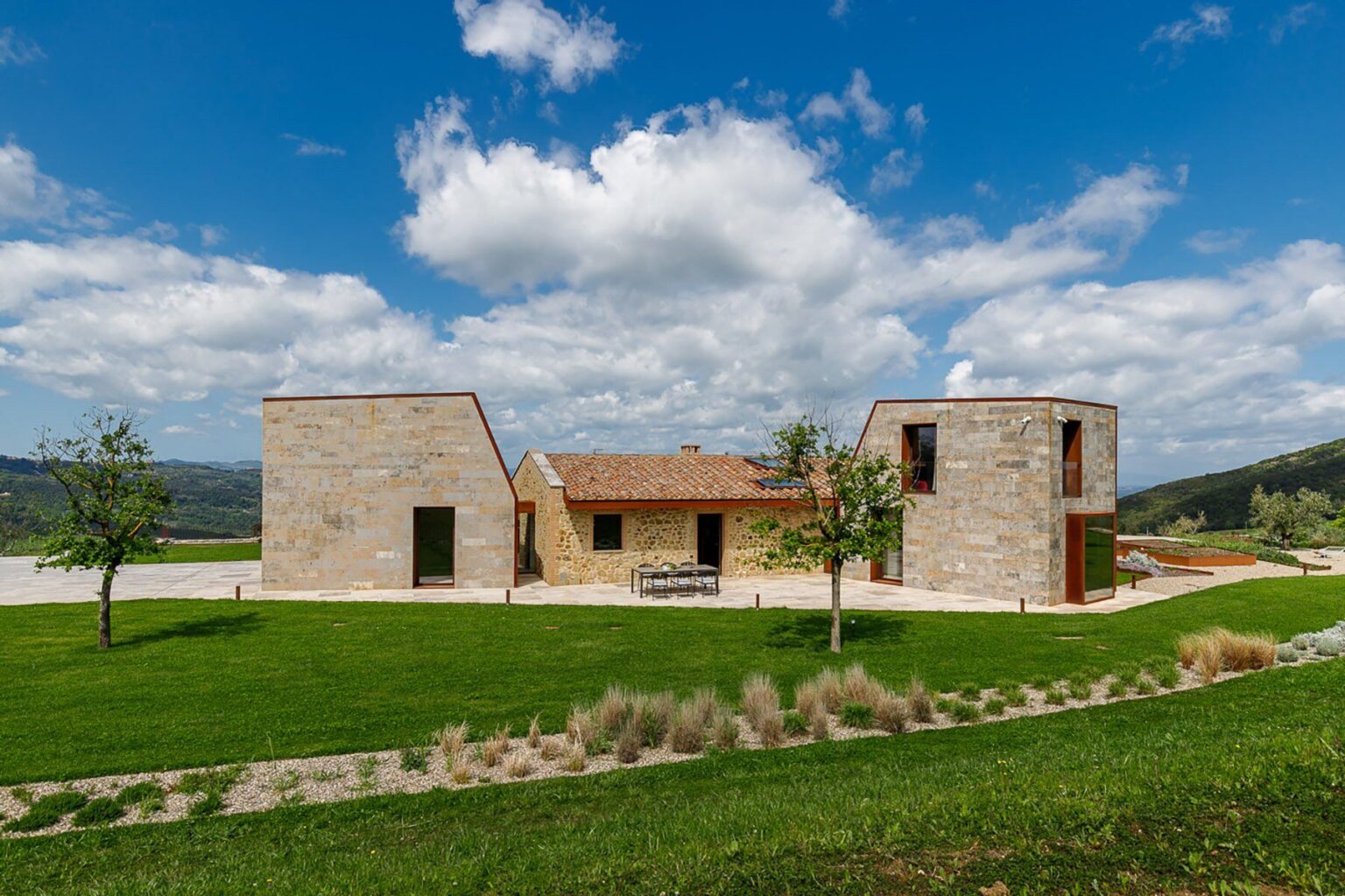 Modern stone house with unique architectural design, surrounded by green lawn and trees, under a blue sky with fluffy clouds. Ideal for showcasing contemporary residential architecture and landscape integration. | Only on SopranoVillas