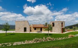 Modern stone house with unique architectural design, surrounded by green lawn and trees, under a blue sky with fluffy clouds. Ideal for showcasing contemporary residential architecture and landscape integration.