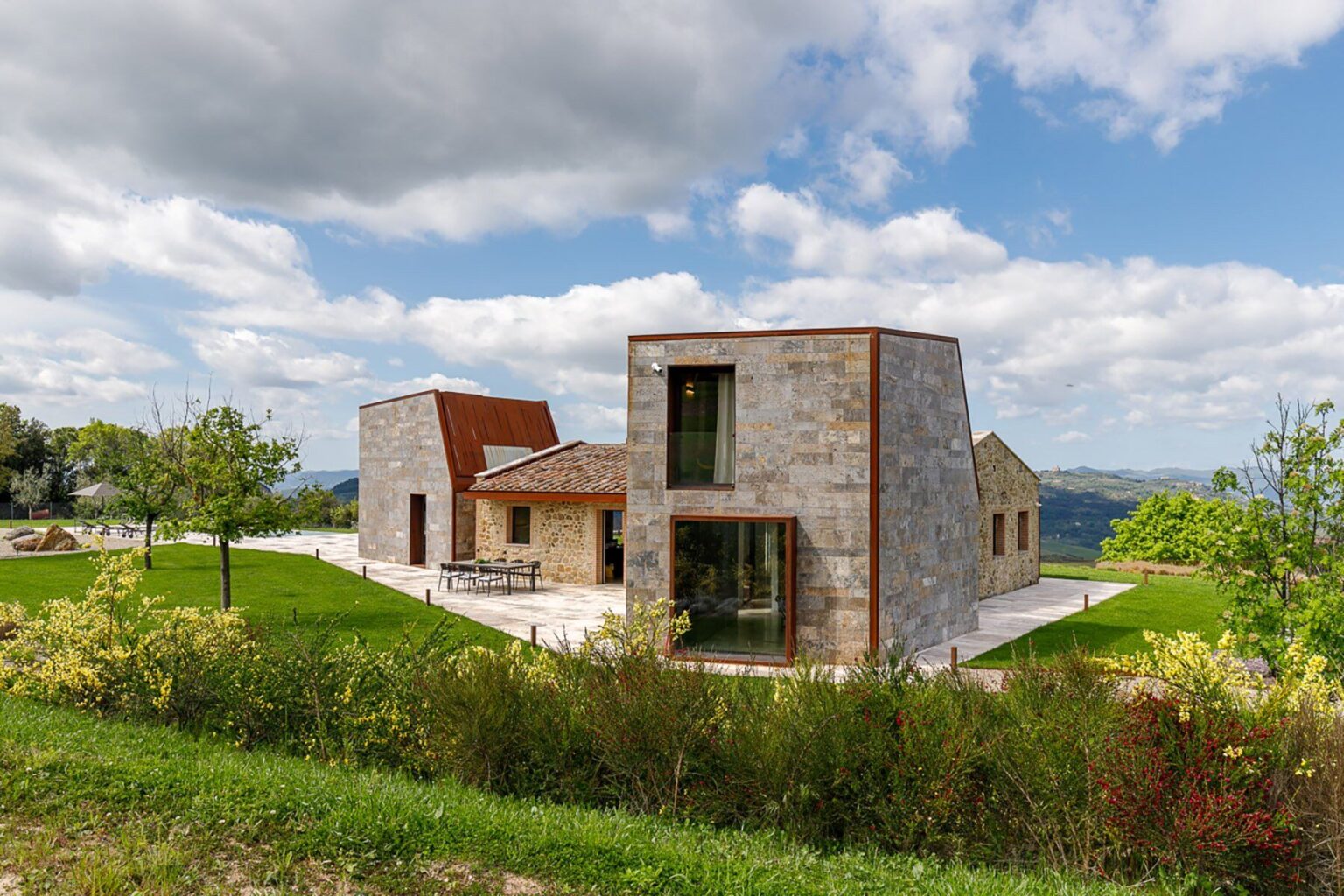 Modern stone house with a unique architectural design, surrounded by lush greenery and under a cloudy sky. The structure features large windows and a combination of stone and metal materials, blending contemporary style with a natural landscape. | Only on SopranoVillas