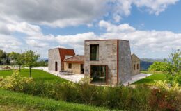 Modern stone house with a unique architectural design, surrounded by lush greenery and under a cloudy sky. The structure features large windows and a combination of stone and metal materials, blending contemporary style with a natural landscape.
