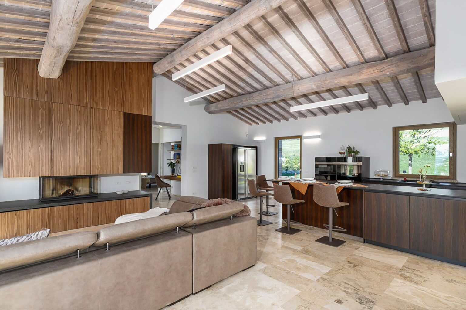 Modern open-concept living space featuring a cozy seating area with a brown leather sofa, wooden ceiling beams, and a stylish kitchen with bar stools. Large windows provide natural light, highlighting the elegant design and warm color palette. | Only on SopranoVillas