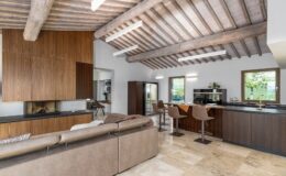 Modern open-concept living space featuring a cozy seating area with a brown leather sofa, wooden ceiling beams, and a stylish kitchen with bar stools. Large windows provide natural light, highlighting the elegant design and warm color palette.