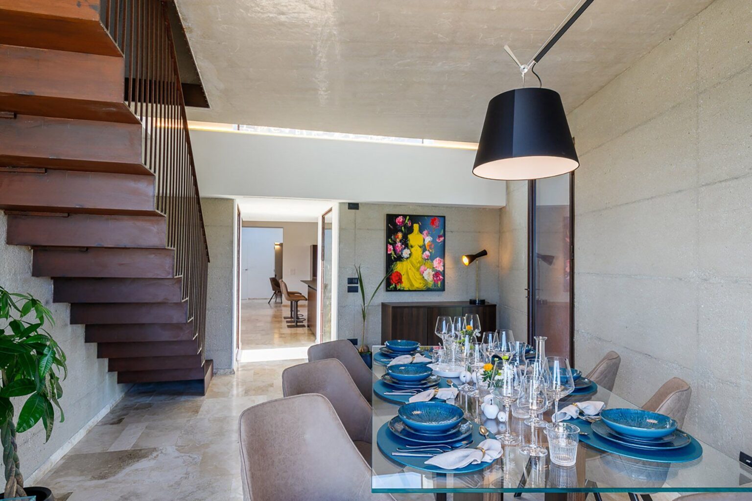 Modern dining room with a glass table set for a meal, featuring blue plates and elegant tableware. A stylish pendant light hangs above, and a staircase made of wood leads to the upper level. The room is bright and airy, showcasing contemporary decor and a colorful artwork on the wall. | Only on SopranoVillas
