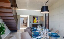 Modern dining room with a glass table set for a meal, featuring blue plates and elegant tableware. A stylish pendant light hangs above, and a staircase made of wood leads to the upper level. The room is bright and airy, showcasing contemporary decor and a colorful artwork on the wall.