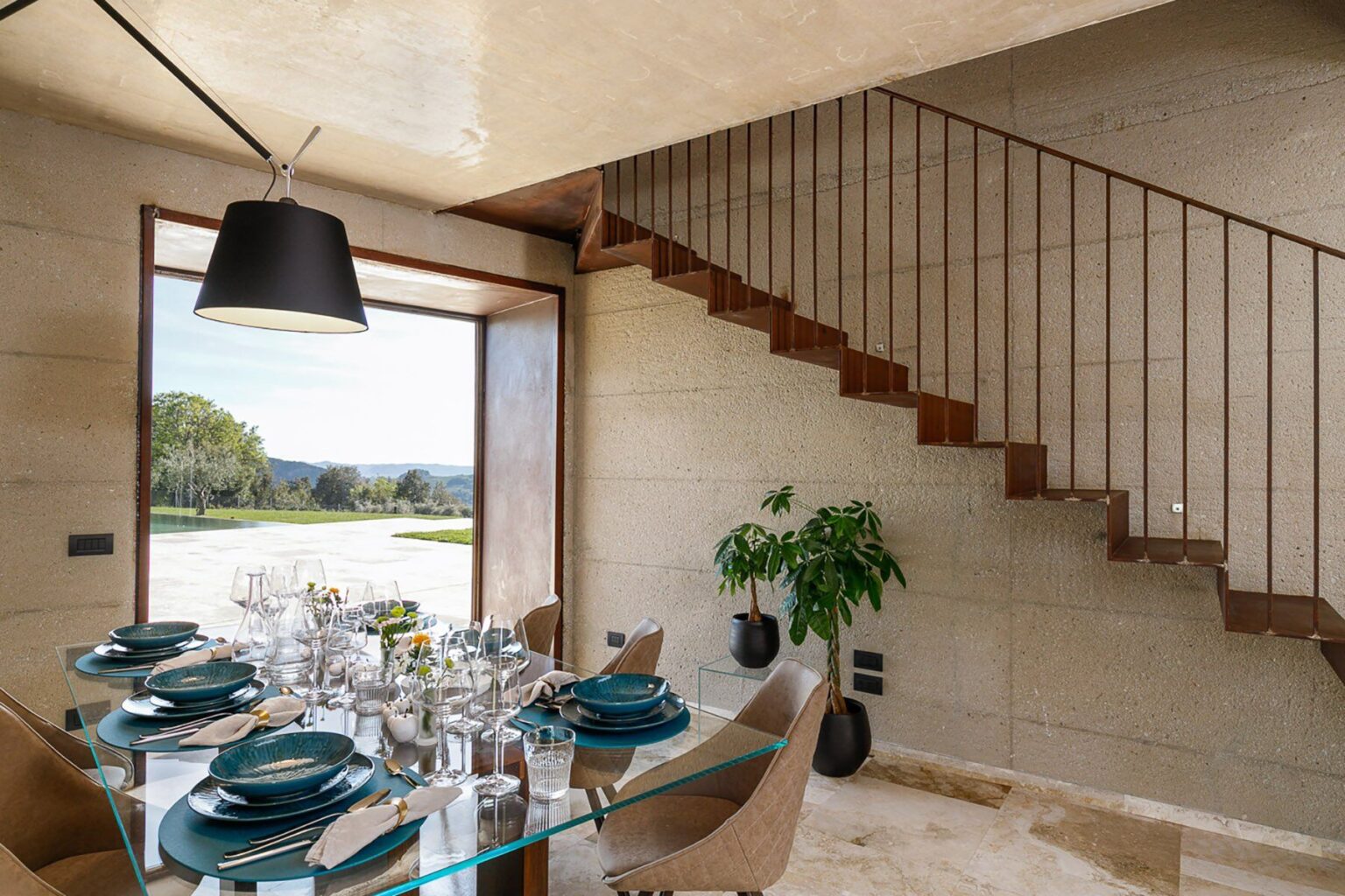 Modern dining area featuring a glass table set with teal dinnerware, surrounded by elegant chairs. A staircase with wooden steps is visible in the background, along with a large window offering a scenic view of the outdoors and a green plant accentuating the space. | Only on SopranoVillas