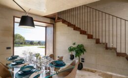 Modern dining area featuring a glass table set with teal dinnerware, surrounded by elegant chairs. A staircase with wooden steps is visible in the background, along with a large window offering a scenic view of the outdoors and a green plant accentuating the space.