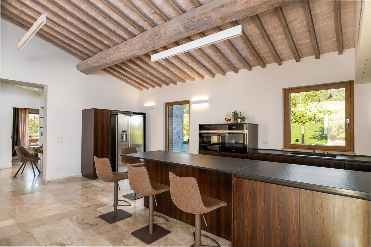 Modern kitchen interior featuring wooden beams, sleek cabinetry, and a spacious island with stylish bar stools. Natural light floods the room through large windows, enhancing the contemporary design and inviting atmosphere. | Only on SopranoVillas