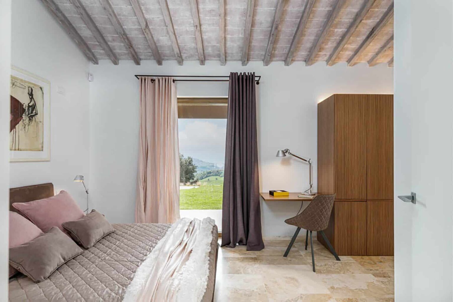 Modern bedroom interior featuring a cozy bed with pink pillows, large windows with curtains, a stylish desk with a lamp, and a wooden wardrobe, all set against a backdrop of green hills. | Only on SopranoVillas