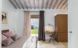 Modern bedroom interior featuring a cozy bed with pink pillows, large windows with curtains, a stylish desk with a lamp, and a wooden wardrobe, all set against a backdrop of green hills.