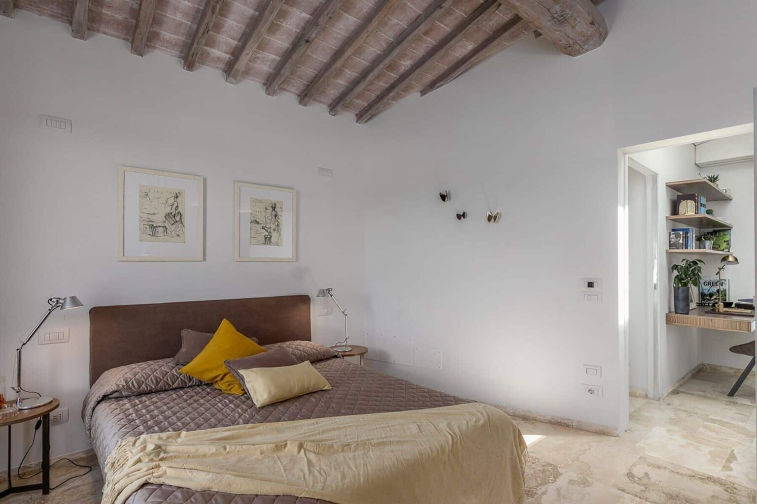 Modern bedroom with wooden beams, featuring a cozy bed adorned with decorative pillows and a soft throw. The light-colored walls and natural light create a bright, inviting atmosphere, complemented by stylish artwork and a small side table. A doorway leads to a well-organized workspace with greenery. | Only on SopranoVillas