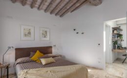 Modern bedroom with wooden beams, featuring a cozy bed adorned with decorative pillows and a soft throw. The light-colored walls and natural light create a bright, inviting atmosphere, complemented by stylish artwork and a small side table. A doorway leads to a well-organized workspace with greenery.