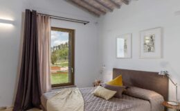 Cozy bedroom featuring a comfortable bed with decorative pillows, large window with scenic views, and elegant curtains. The room is adorned with framed artwork and has a modern aesthetic with wooden beams on the ceiling. Ideal for relaxation and comfort.