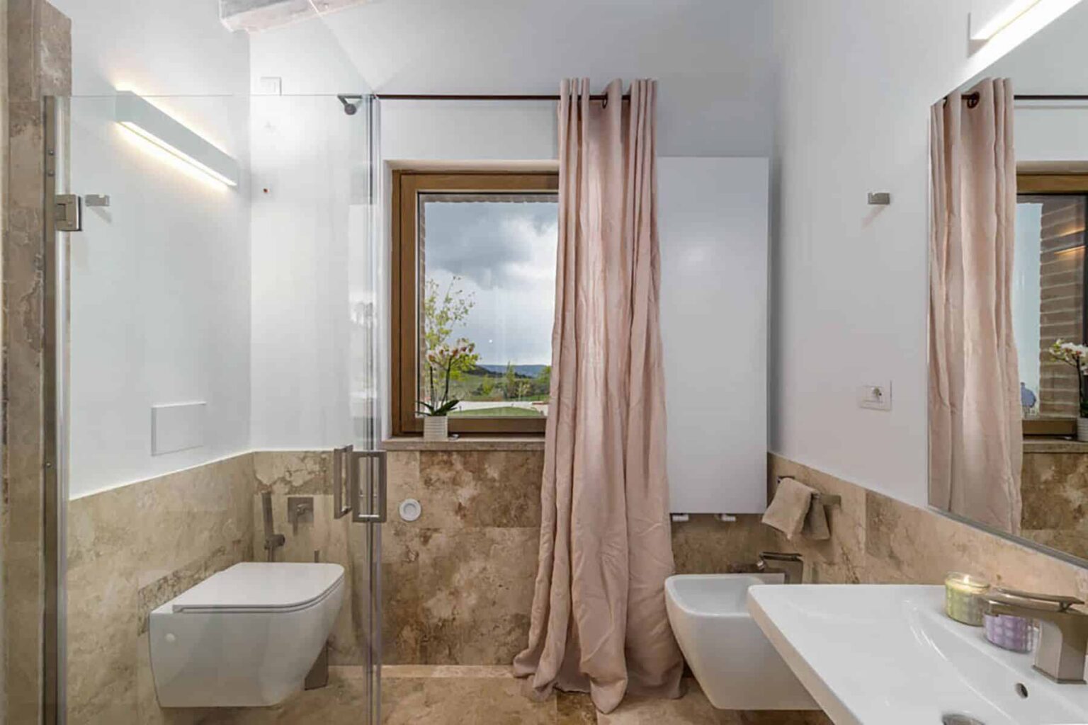 Modern bathroom featuring a walk-in shower with glass doors, a sleek toilet, and a stylish sink. Natural light streams in through a window adorned with soft pink curtains, highlighting the elegant stone tiles and minimalist design. | Only on SopranoVillas