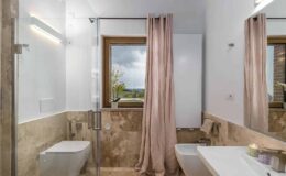 Modern bathroom featuring a walk-in shower with glass doors, a sleek toilet, and a stylish sink. Natural light streams in through a window adorned with soft pink curtains, highlighting the elegant stone tiles and minimalist design.