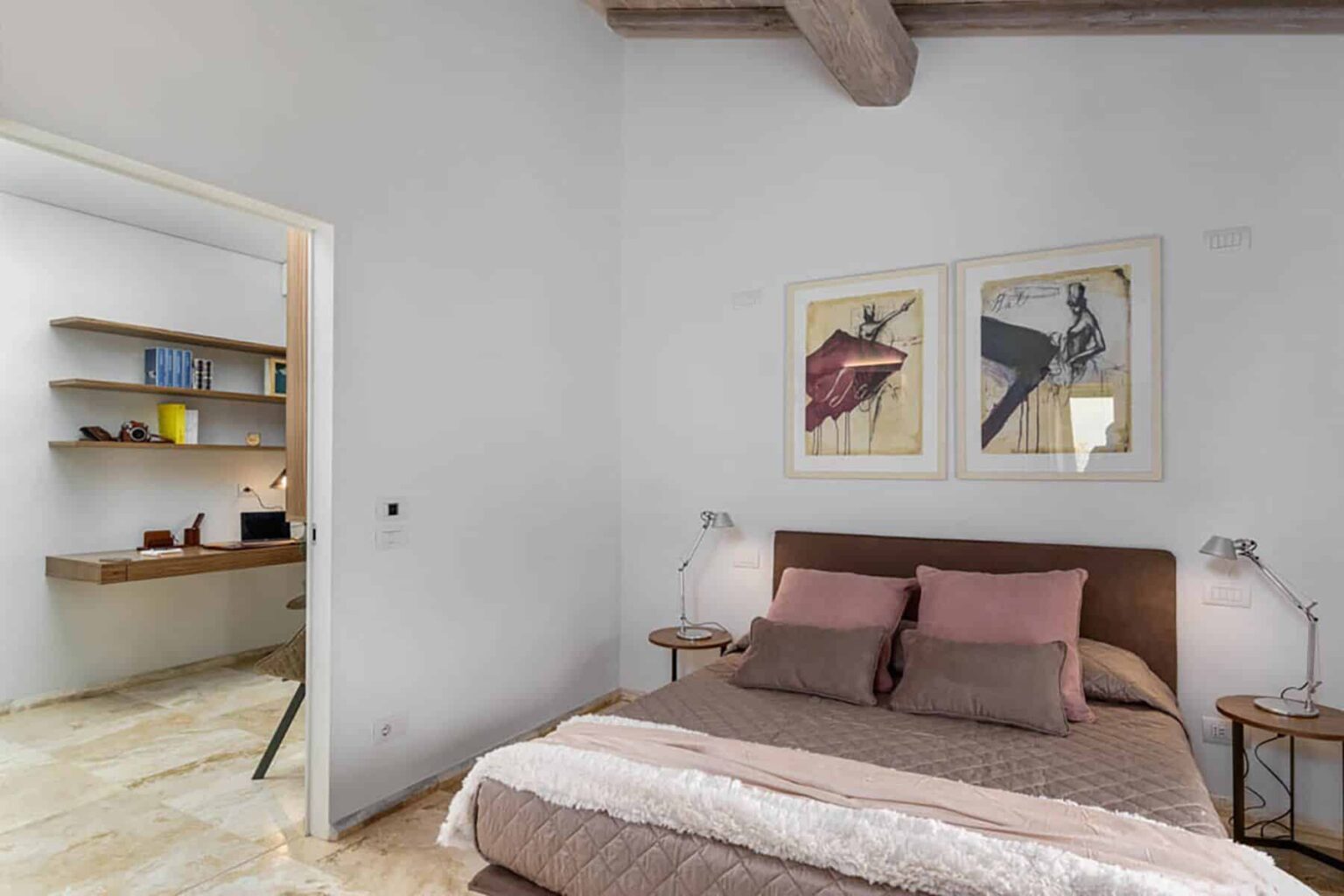 Cozy bedroom featuring a comfortable bed with pink and gray bedding, framed artwork on the walls, and modern bedside lamps. A stylish workspace is visible in the adjacent room, highlighting a minimalist design with wooden shelves. | Only on SopranoVillas