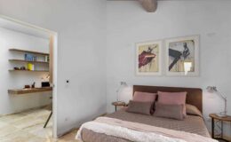 Cozy bedroom featuring a comfortable bed with pink and gray bedding, framed artwork on the walls, and modern bedside lamps. A stylish workspace is visible in the adjacent room, highlighting a minimalist design with wooden shelves.