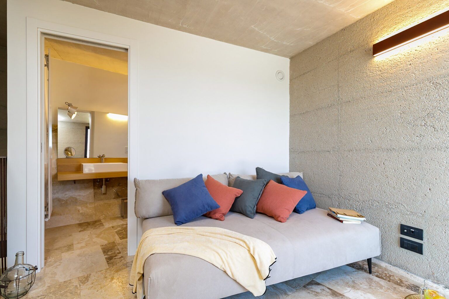 Cozy living space featuring a modern sofa adorned with colorful throw pillows, a soft blanket, and a warm lighting fixture. The room opens to a stylish bathroom area, showcasing contemporary design and neutral tones. Ideal for relaxation and comfort. | Only on SopranoVillas