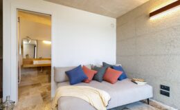 Cozy living space featuring a modern sofa adorned with colorful throw pillows, a soft blanket, and a warm lighting fixture. The room opens to a stylish bathroom area, showcasing contemporary design and neutral tones. Ideal for relaxation and comfort.