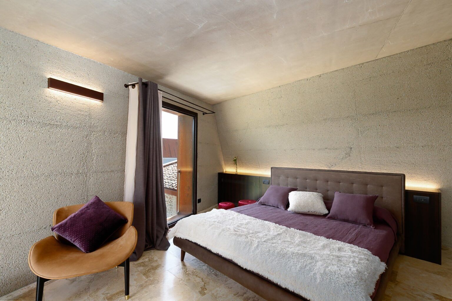 Modern bedroom design featuring a plush bed with purple and white bedding, a stylish chair, and large windows allowing natural light. The walls are finished in a textured concrete, creating a contemporary aesthetic. | Only on SopranoVillas