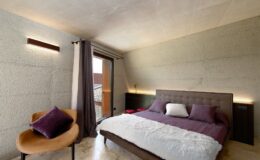 Modern bedroom design featuring a plush bed with purple and white bedding, a stylish chair, and large windows allowing natural light. The walls are finished in a textured concrete, creating a contemporary aesthetic.