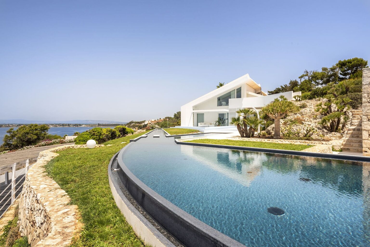 Modern coastal villa with an infinity pool overlooking the ocean, surrounded by lush greenery and rocky terrain under a clear blue sky. | Only on SopranoVillas