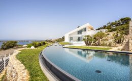 Modern coastal villa with an infinity pool overlooking the ocean, surrounded by lush greenery and rocky terrain under a clear blue sky.