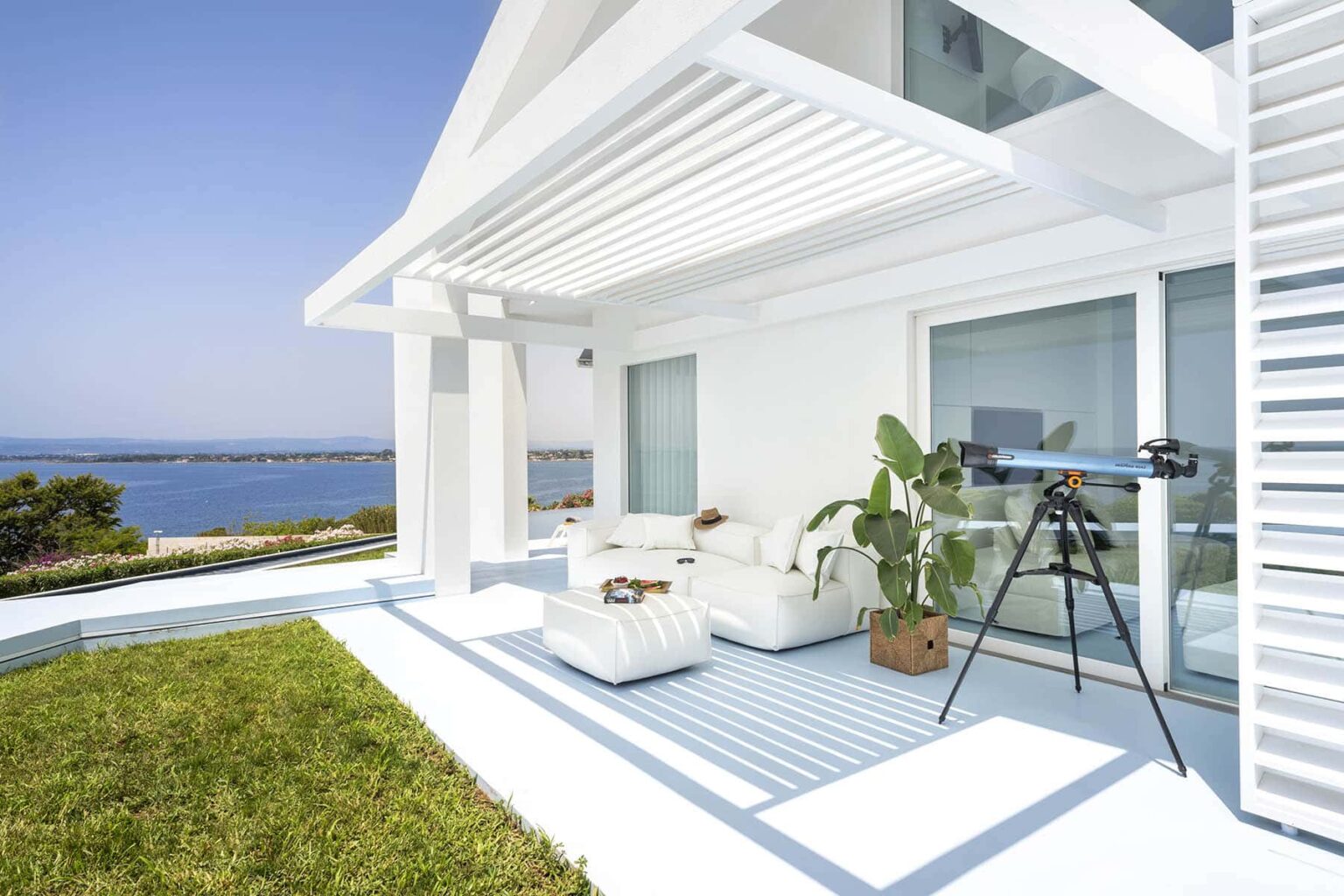 Modern coastal patio with white furniture, a telescope, and ocean views, featuring lush greenery and bright blue skies. Ideal for relaxation and outdoor enjoyment. | Only on SopranoVillas