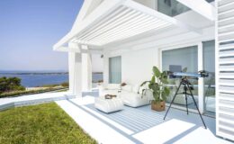 Modern coastal patio with white furniture, a telescope, and ocean views, featuring lush greenery and bright blue skies. Ideal for relaxation and outdoor enjoyment.