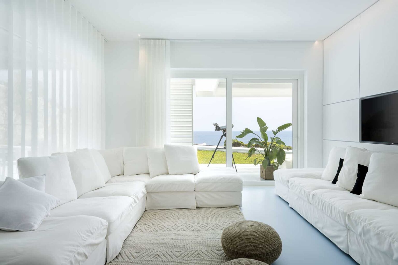 Modern living room with a large white sectional sofa, minimalist decor, and large windows offering a view of the ocean. A potted plant adds a touch of greenery, while natural light enhances the bright, airy atmosphere. | Only on SopranoVillas