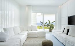 Modern living room with a large white sectional sofa, minimalist decor, and large windows offering a view of the ocean. A potted plant adds a touch of greenery, while natural light enhances the bright, airy atmosphere.